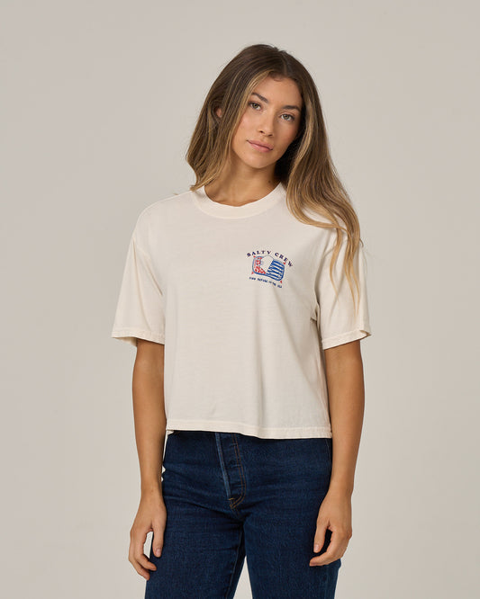 A woman stands against a plain light background, looking at the camera with a neutral expression, wearing the Salty Crew Cute Tin Skimmer Tee in Off White—a boxy style featuring a small screen print graphic and text on the left chest.