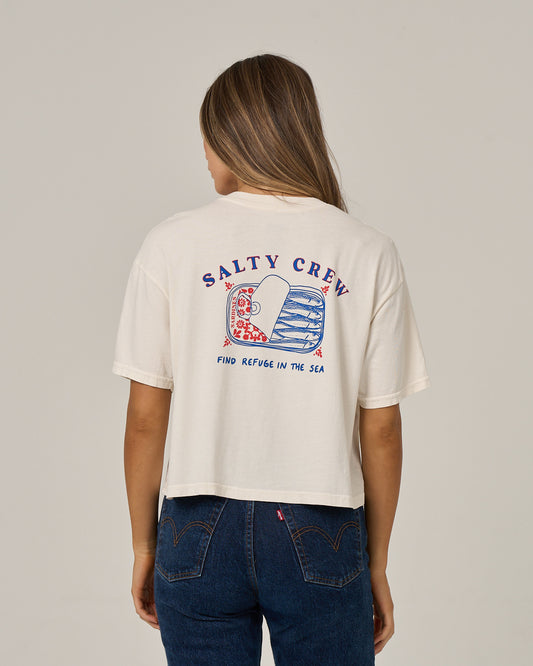 A woman wearing the Salty Crew Cute Tin Skimmer Tee in off white—a cotton jersey shirt featuring a fish graphic and the “Find Refuge in the Sea” slogan screen printed on the back—paired with blue jeans, standing against a plain background.