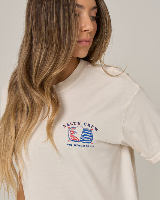 A woman with long, wavy, light brown hair wears the Salty Crew Cute Tin Skimmer Tee in off white, which features small, colorful graphics and “Find refuge in the sea” text on the chest. She is looking to her left.