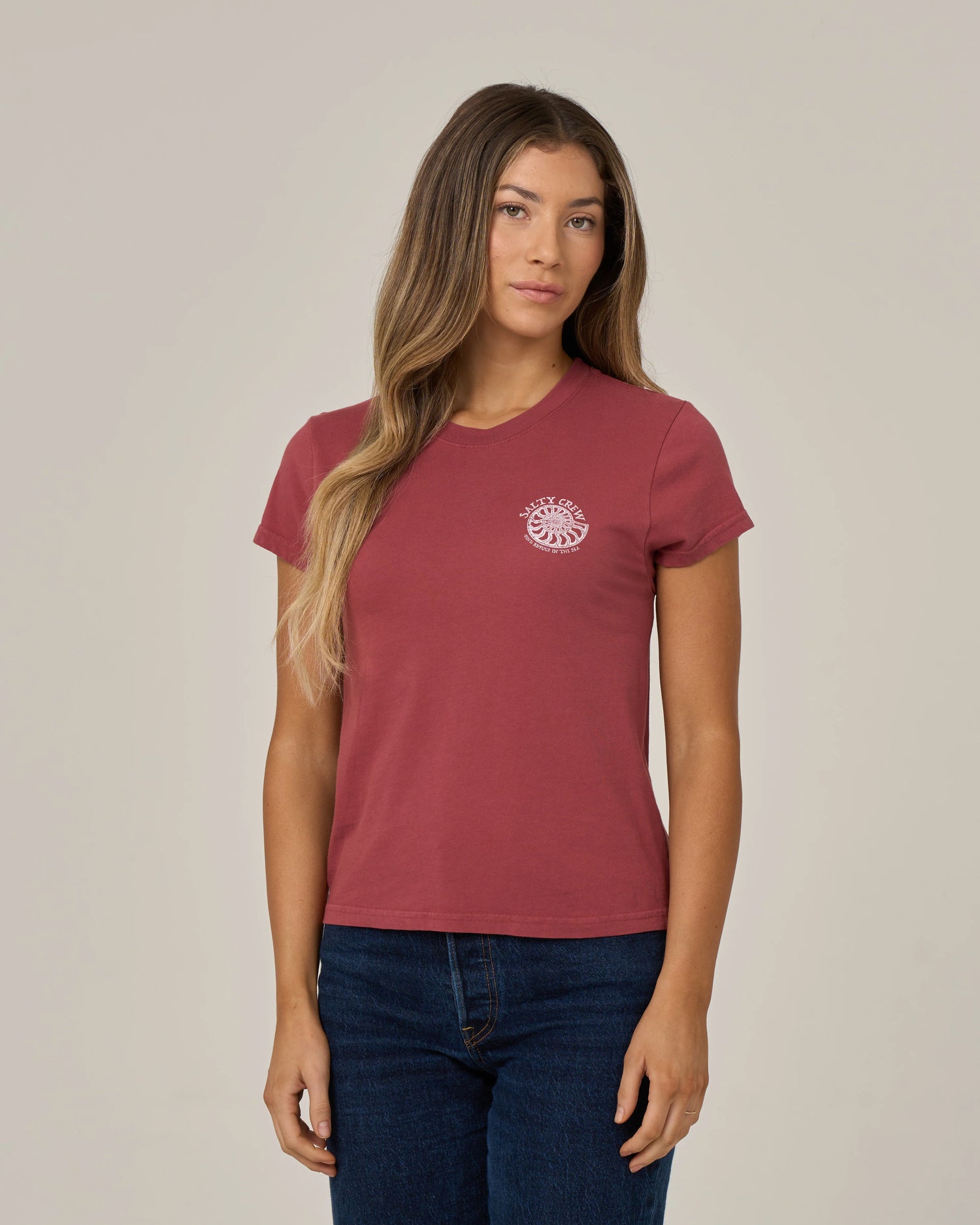 A woman with long, wavy hair stands against a plain, light-colored background, wearing the Salty Crew Nautilus Modern Tee in Baked Apple—a maroon cotton jersey top featuring cap sleeves and a small white sun graphic on the chest.