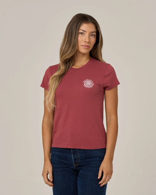 A woman with long, wavy hair stands against a plain, light-colored background, wearing the Salty Crew Nautilus Modern Tee in Baked Apple—a maroon cotton jersey top featuring cap sleeves and a small white sun graphic on the chest.