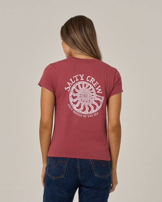 A woman with long hair, wearing the Salty Crew Nautilus Modern Tee in Baked Apple and blue jeans, is facing away. The cotton jersey shirt, designed with cap sleeves, features a white screen print of a nautilus shell and the words “Salty Crew, Find Refuge In The Sea” on the back.