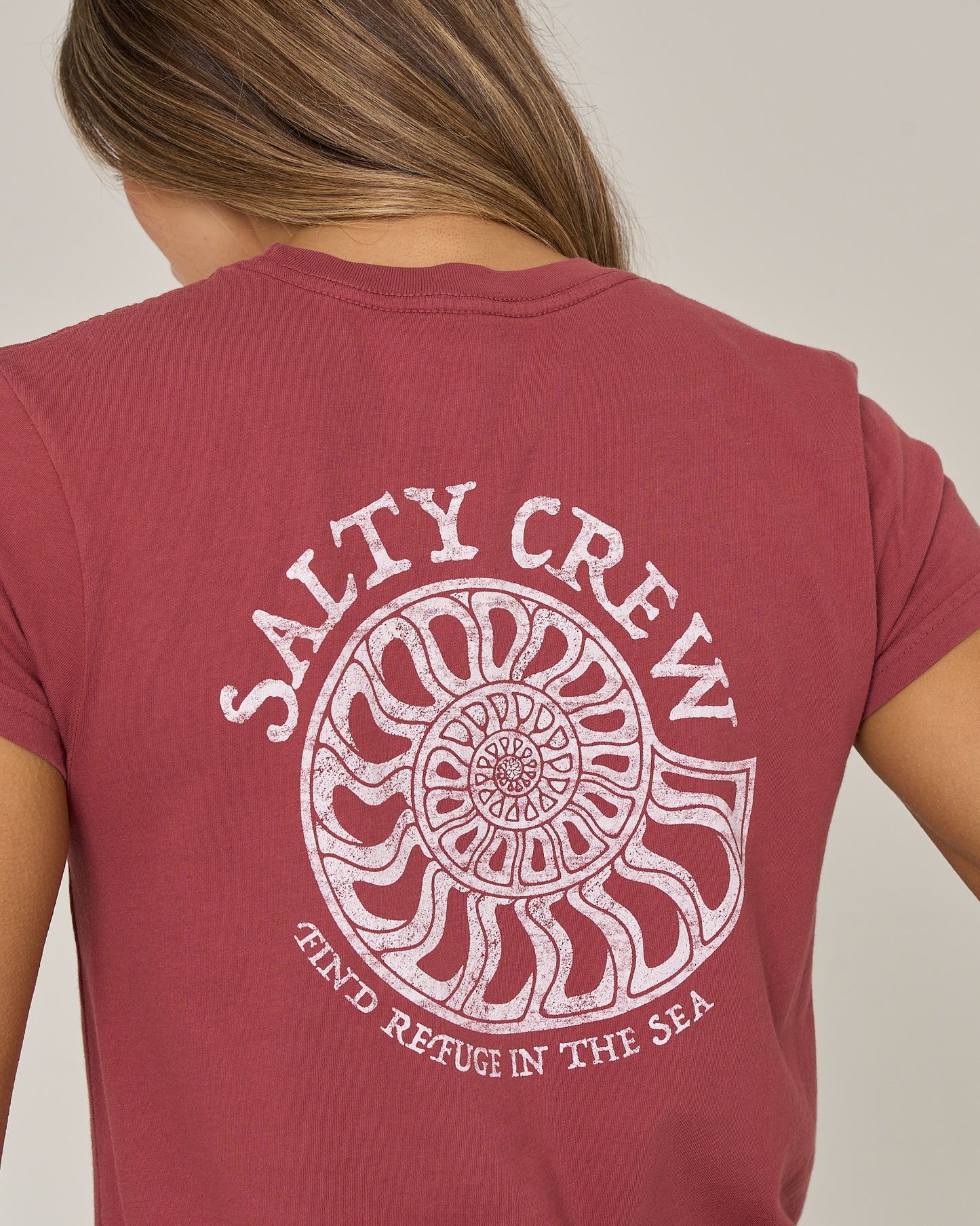 A person wearing the Salty Crew Nautilus Modern Tee in Baked Apple, featuring a white nautilus shell screen print and the words “Salty Crew” and “Find Refuge In The Sea” on the back.