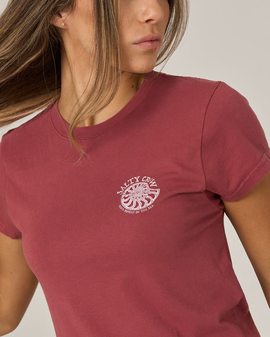 A woman wearing the Salty Crew Nautilus Modern Tee in Baked Apple, featuring a white screen-printed logo on the chest, looks to the side with her brown hair partially covering her face. The background is plain and light.