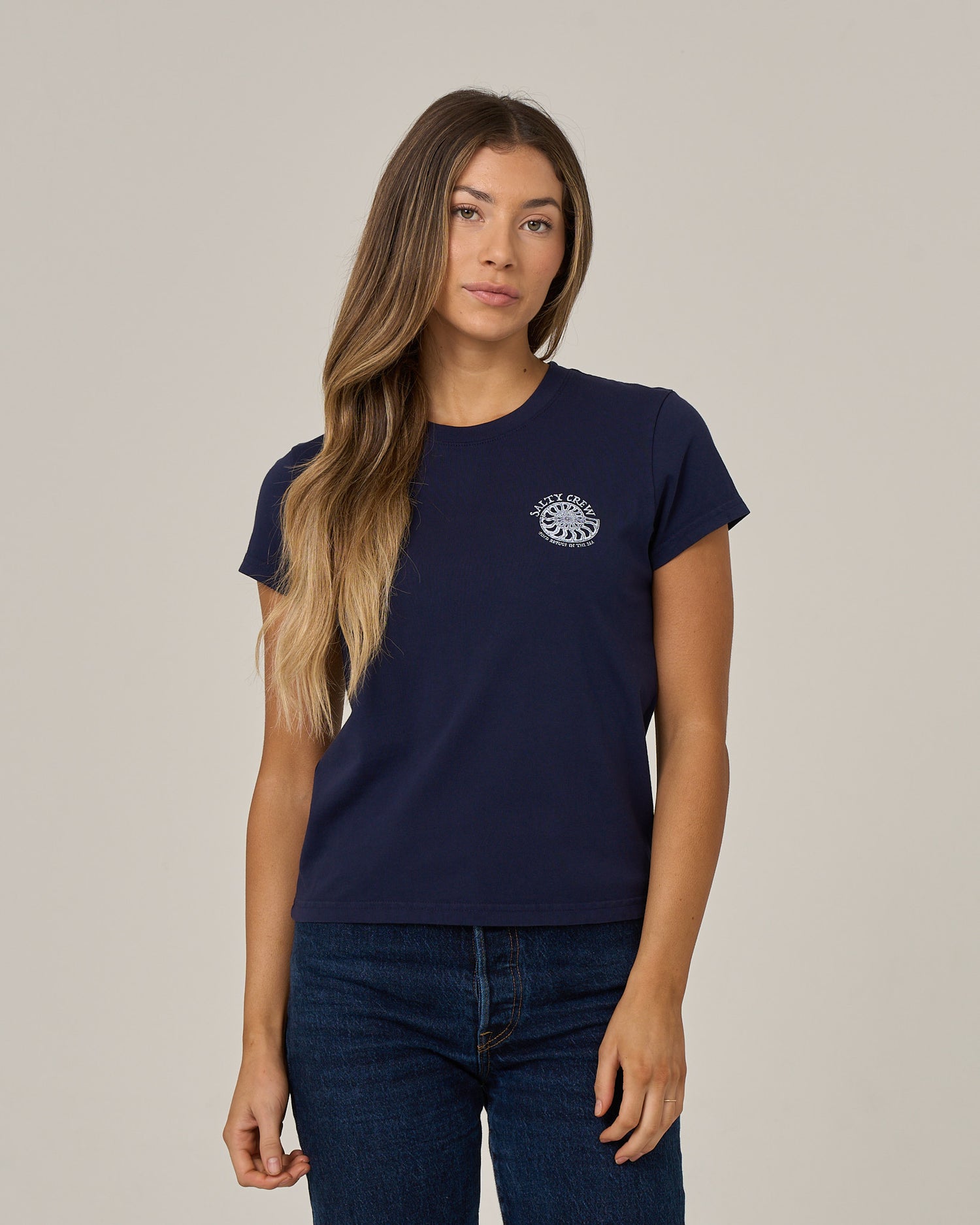 A woman with long, loose hair and a neutral expression stands against a plain light background, wearing the Salty Crew Nautilus Modern Tee in bright navy—a cotton jersey tee with cap sleeves and a small white circular screen print logo on the left chest.