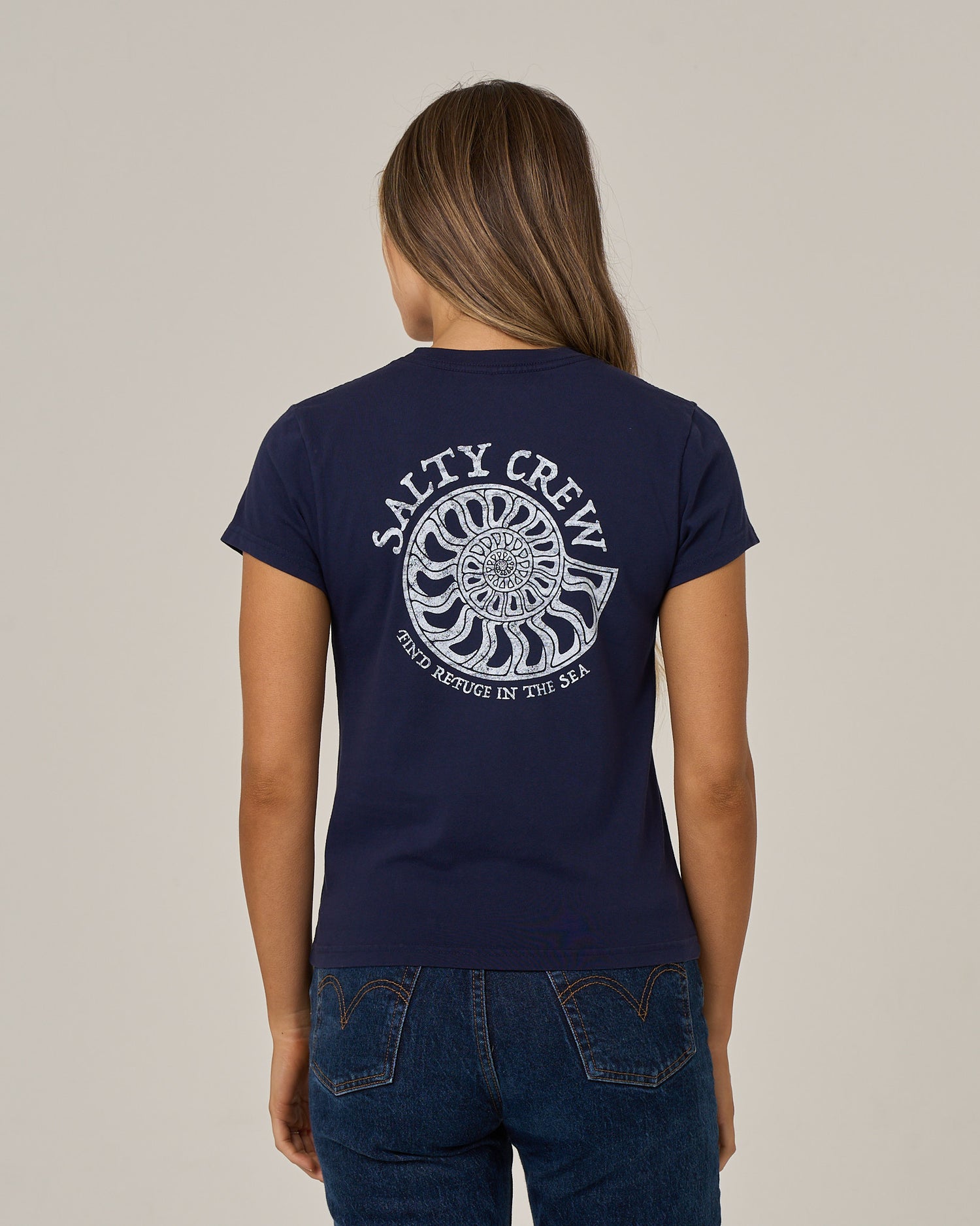 A person with long hair is wearing the Salty Crew Nautilus Modern Tee in Bright Navy, which features a white nautilus shell graphic and the words “Salty Crew Find Refuge in the Sea” on the back, standing against a plain light background.