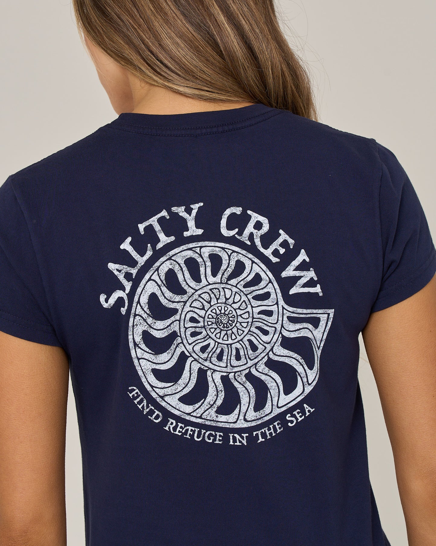 A person wearing the Salty Crew Nautilus Modern Tee in bright navy—a cotton jersey shirt with cap sleeves, featuring a white nautilus shell screen print and the words “Salty Crew” and “Find Refuge in the Sea” on the back.