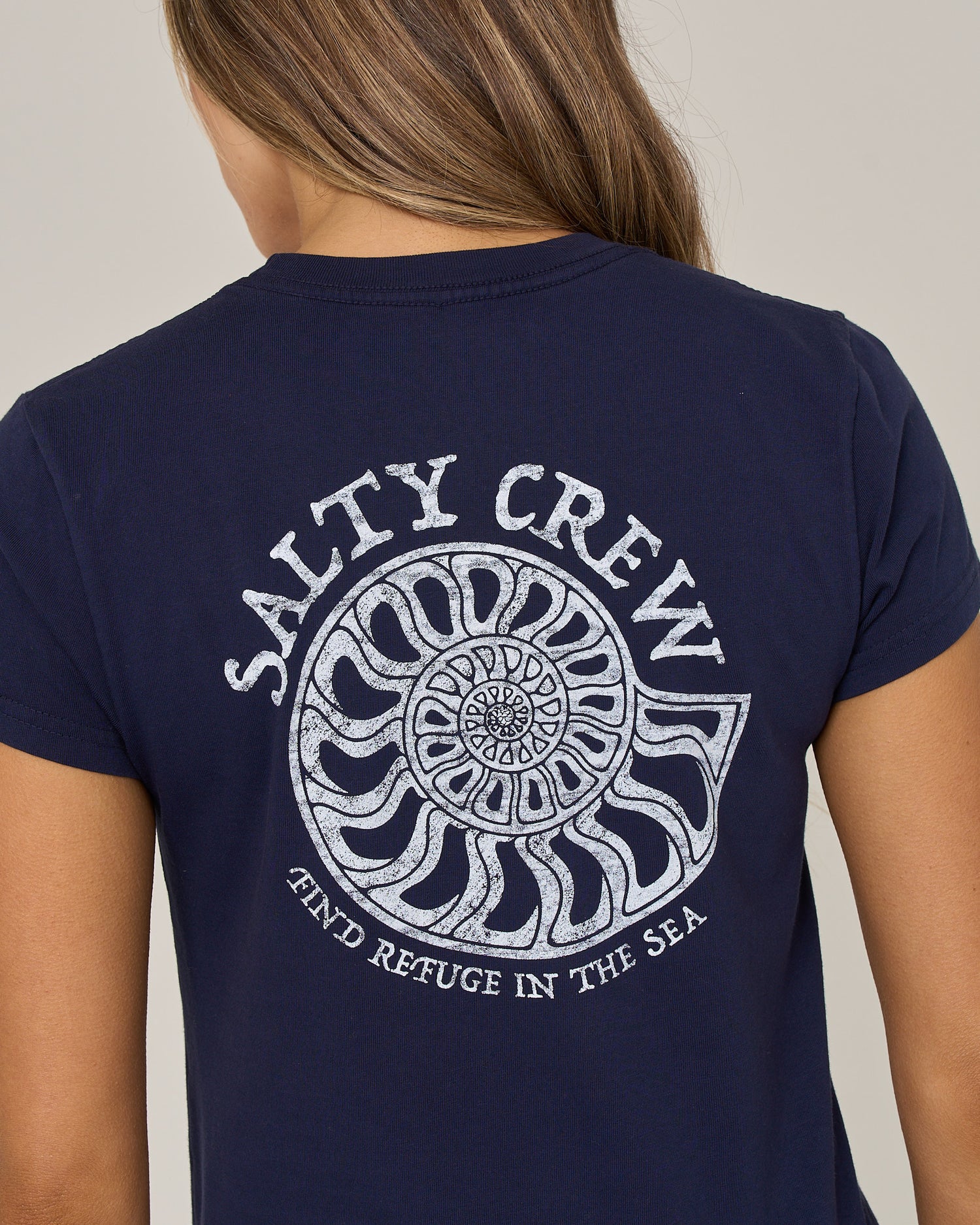 A person wearing the Salty Crew Nautilus Modern Tee in bright navy—a cotton jersey shirt with cap sleeves, featuring a white nautilus shell screen print and the words “Salty Crew” and “Find Refuge in the Sea” on the back.