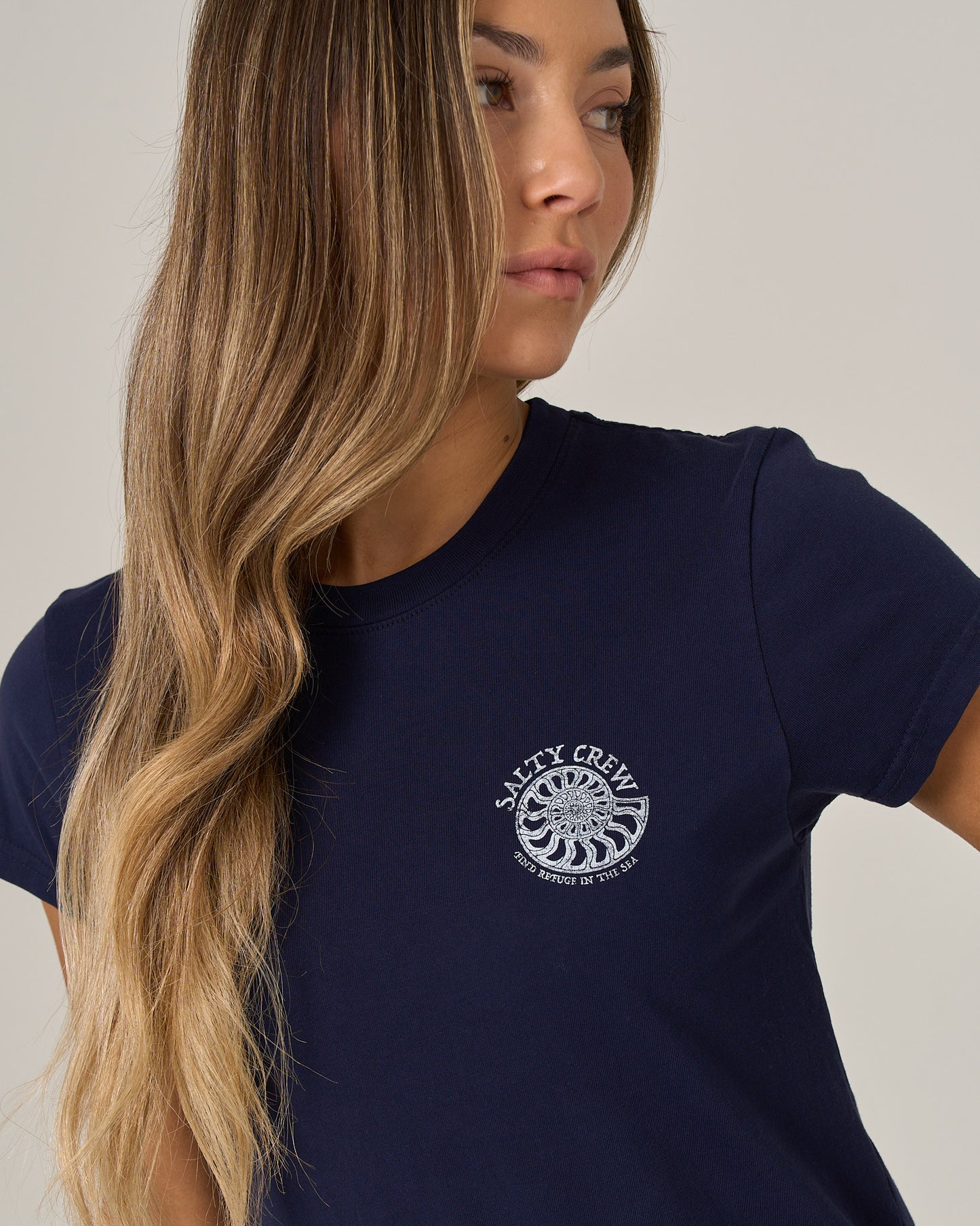 A woman with long wavy blonde hair wears the Salty Crew Nautilus Modern Tee in Bright Navy, featuring cap sleeves, as she looks to the side against a plain background.
