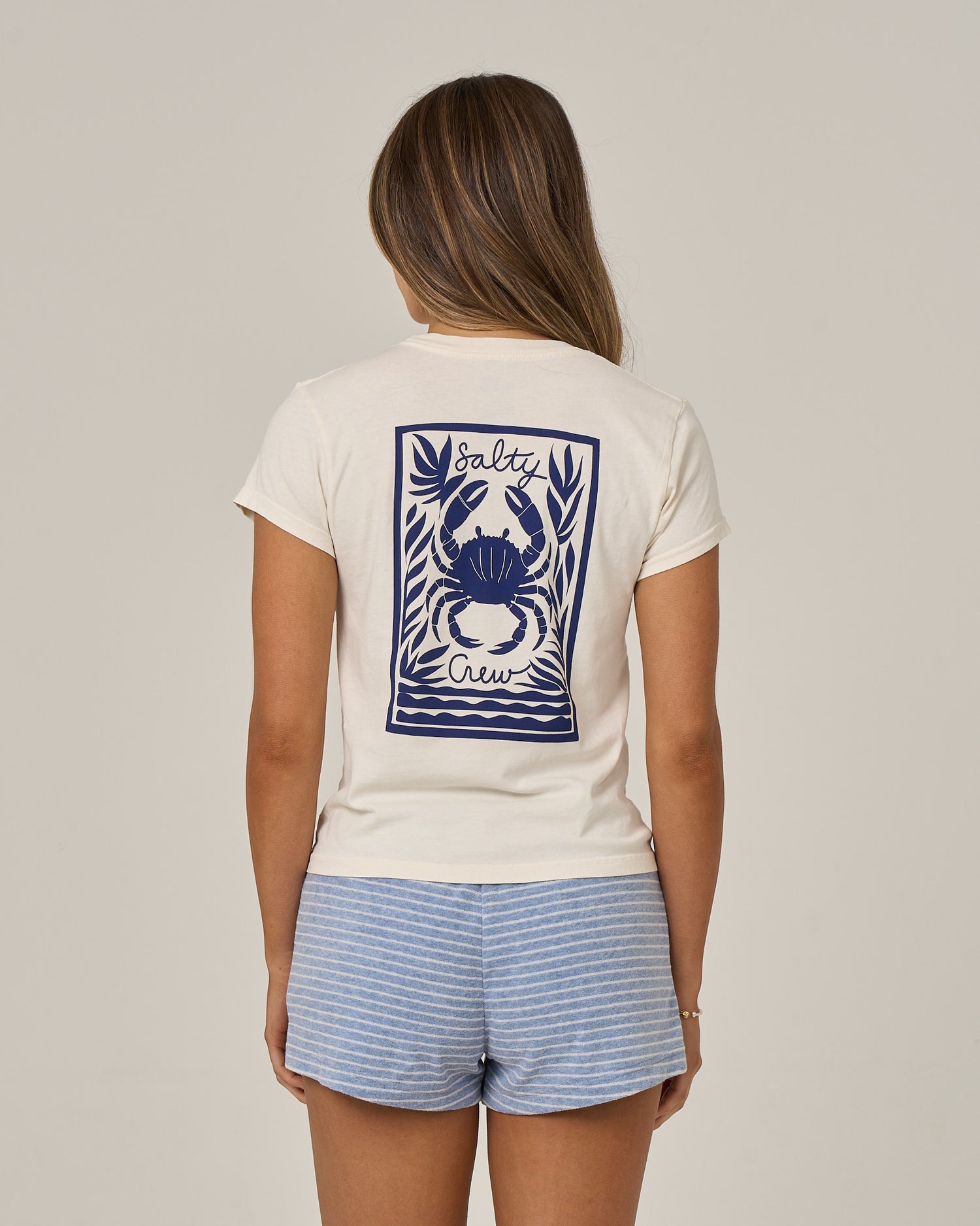 Wearing the Salty Crew Crabby Modern Tee in off white, a woman with long hair stands against a plain background. The cotton jersey tee features a blue crab graphic and Salty Crew text on the back, and is paired with blue and white striped shorts.