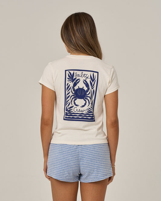 Wearing the Salty Crew Crabby Modern Tee in off white, a woman with long hair stands against a plain background. The cotton jersey tee features a blue crab graphic and Salty Crew text on the back, and is paired with blue and white striped shorts.