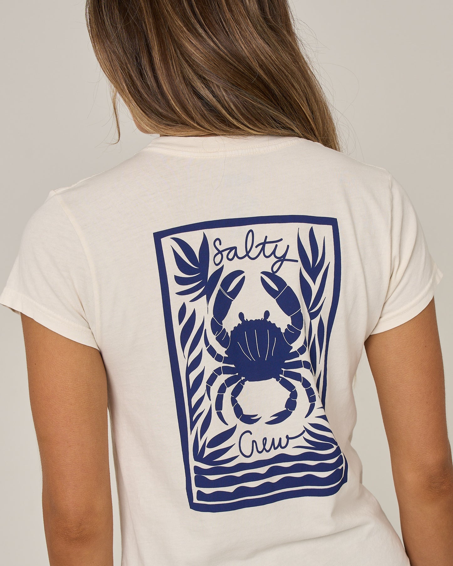A woman wearing the Salty Crew Crabby Modern Tee in off white—a cotton jersey t-shirt with cap sleeves, featuring a blue crab and seaweed graphic along with “Salty Crew” printed on the back.