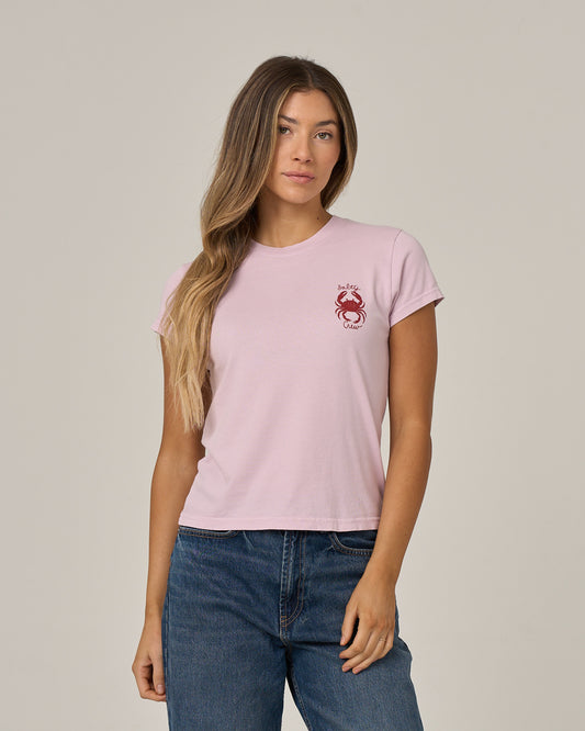 A woman stands against a plain background, looking at the camera with one hand by her side and the other in her pocket, wearing the Salty Crew Crabby Modern Tee in Pale Orchid—a light pink fitted top featuring cap sleeves and a small red crab graphic on the chest.