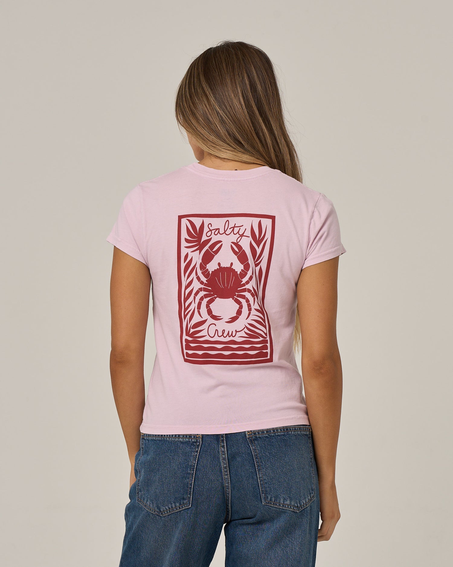 A woman with long hair is seen from behind wearing the Salty Crew Crabby Modern Tee in Pale Orchid, a light pink cotton jersey shirt with cap sleeves, featuring a red crab graphic and Salty Crew text on the back, paired with blue jeans.