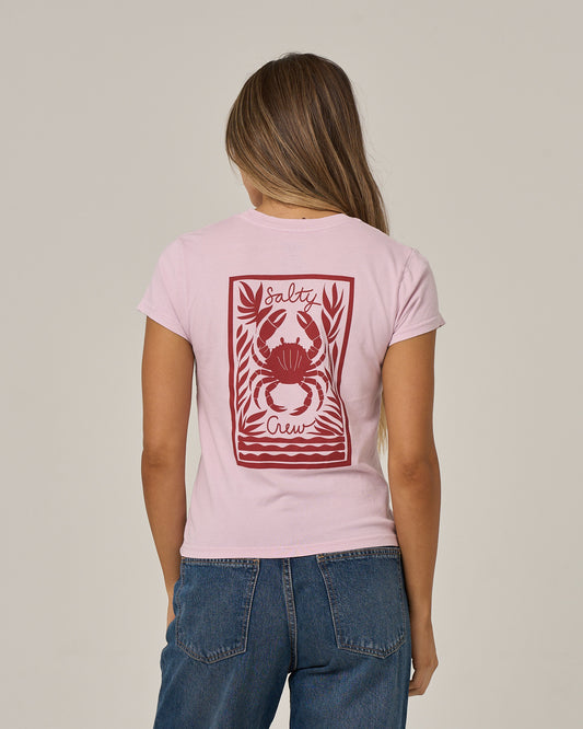 A woman with long hair is seen from behind wearing the Salty Crew Crabby Modern Tee in Pale Orchid, a light pink cotton jersey shirt with cap sleeves, featuring a red crab graphic and Salty Crew text on the back, paired with blue jeans.