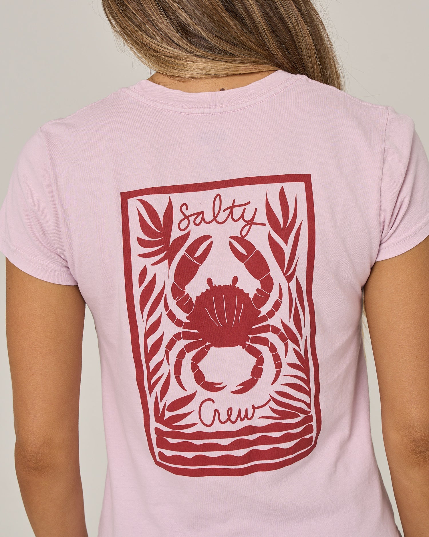 A person is shown from behind wearing the Salty Crew Crabby Modern Tee in Pale Orchid, a fitted cotton jersey shirt featuring a red graphic of a crab and seaweed with “Salty Crew” on the back.