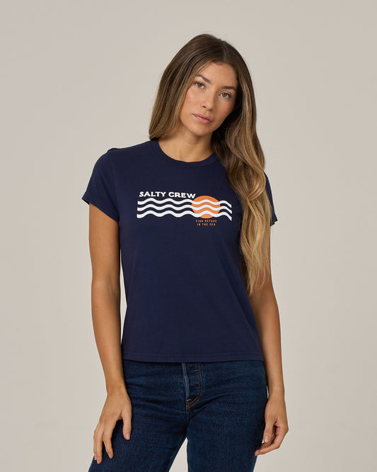 A woman with long, wavy hair wears the Salty Crew Boat Legs Modern Tee in bright navy—a fitted style crafted from soft cotton jersey, featuring white and orange wave graphics and cap sleeves—and pairs it with dark jeans as she stands against a plain light background.