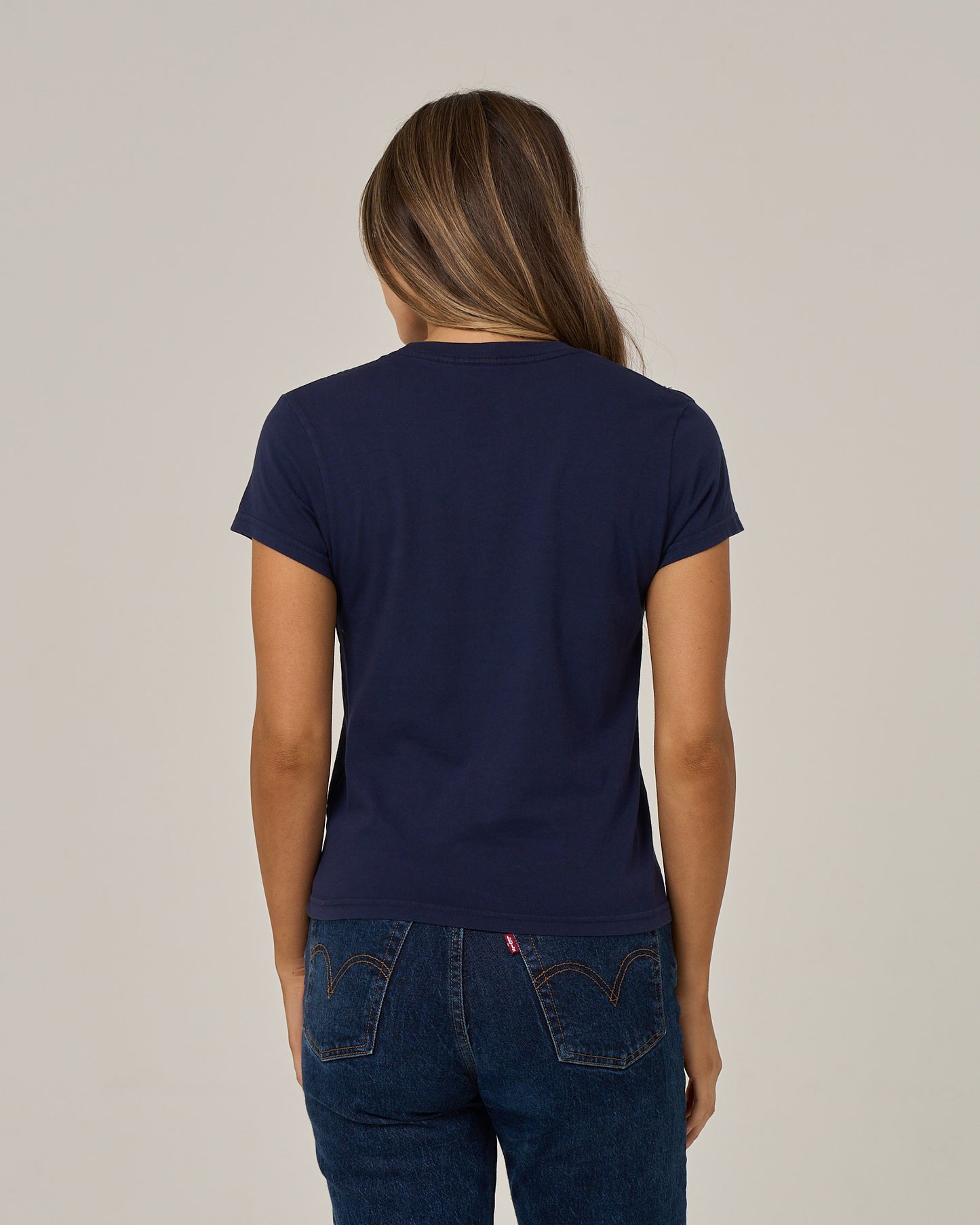 A person with long hair is seen from the back, standing against a plain light background and wearing the Salty Crew Boat Legs Modern Tee in Bright Navy paired with blue jeans.