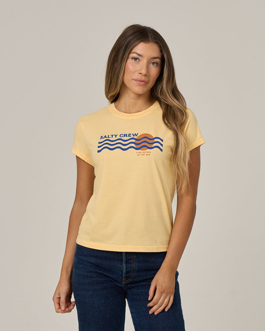 A woman with long hair wears the Salty Crew Boat Legs Modern Tee in Custard, featuring wave graphics, paired with blue jeans and standing against a plain light background.