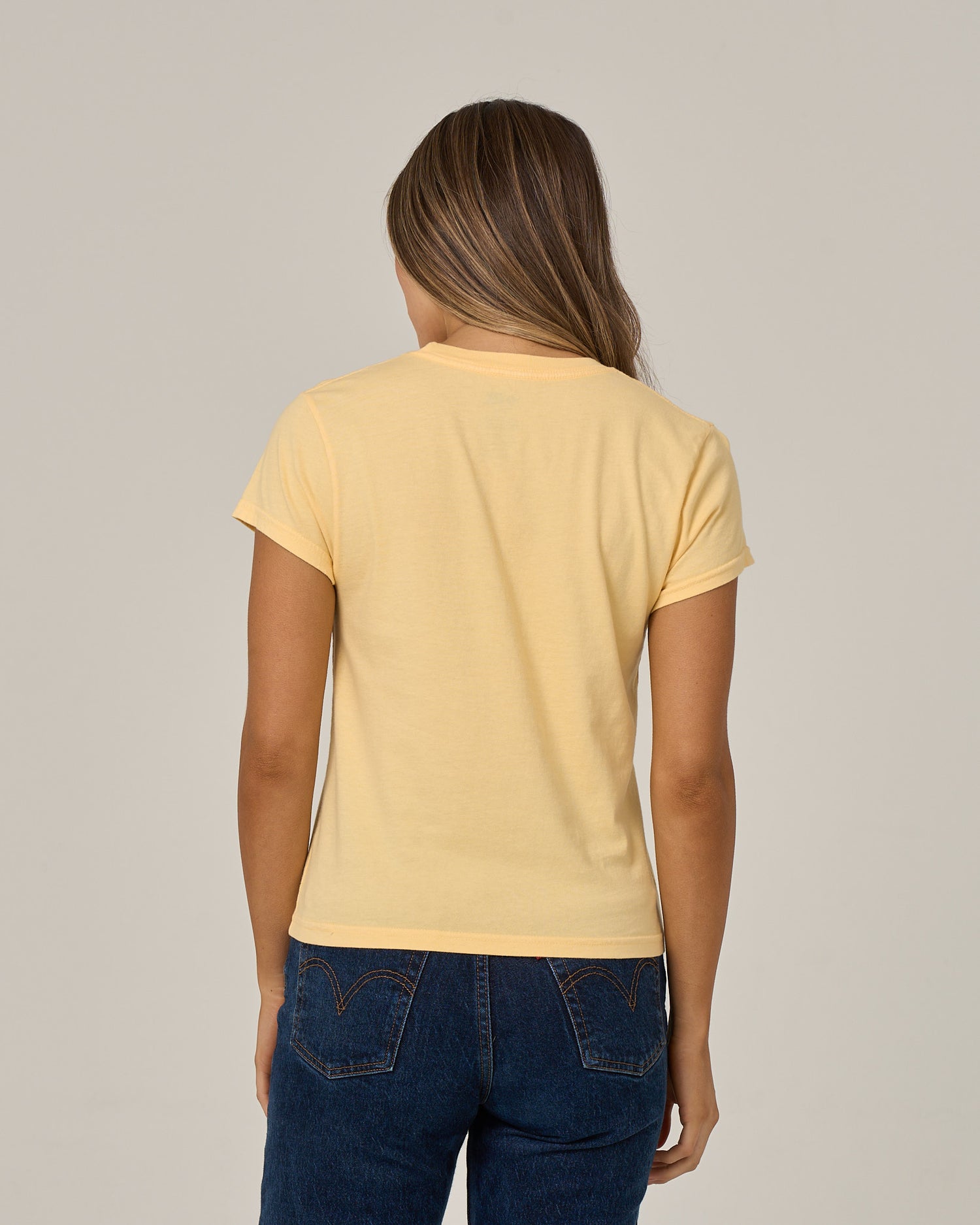 A person with long brown hair, wearing the Salty Crew Boat Legs Modern Tee in Custard—a fitted yellow cotton jersey t-shirt with cap sleeves—paired with blue jeans, is standing facing away from the camera against a plain light background.