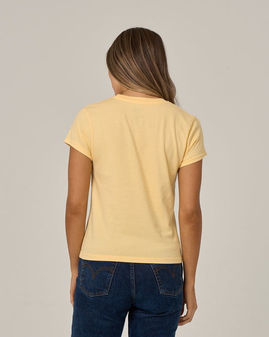 A person with long brown hair, wearing the Salty Crew Boat Legs Modern Tee in Custard—a fitted yellow cotton jersey t-shirt with cap sleeves—paired with blue jeans, is standing facing away from the camera against a plain light background.