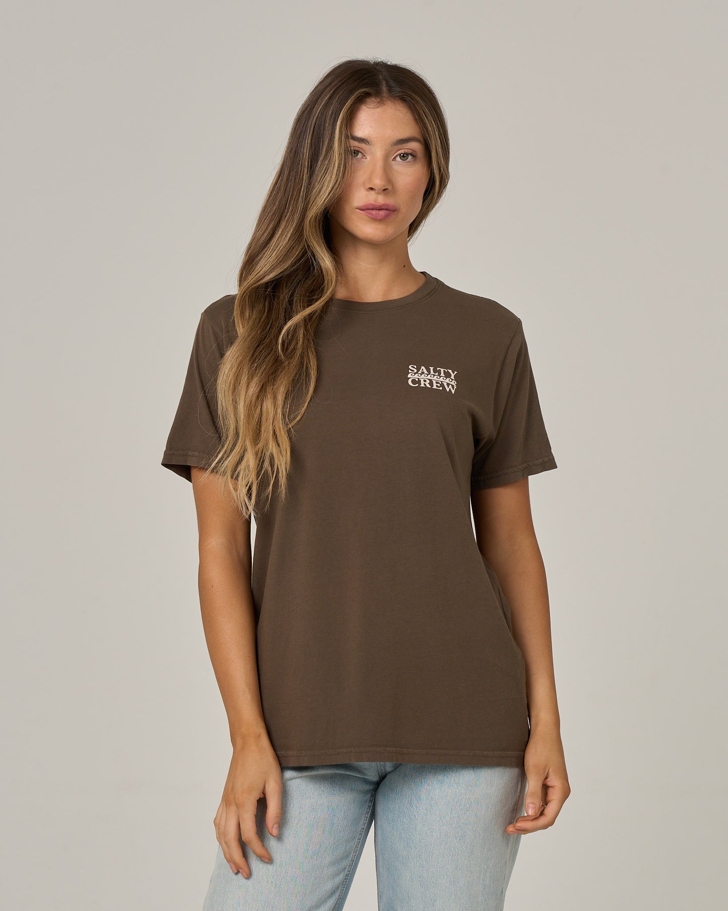 A woman with long, wavy hair wears the Salty Crew Lobster Roll Boyfriend Tee in washed brown and light blue jeans, standing against a plain, light background.