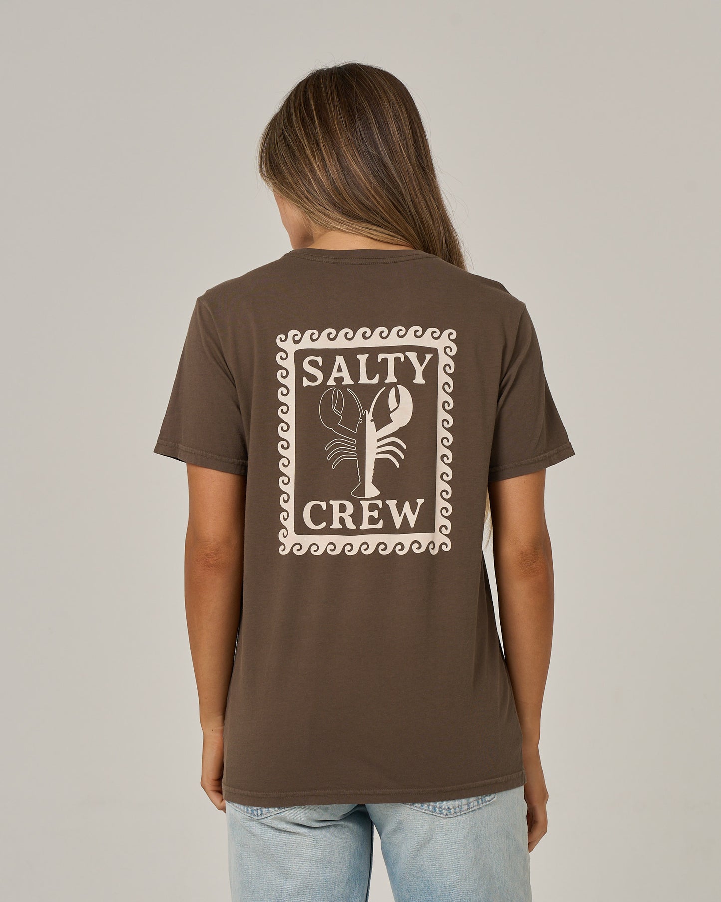 A person stands with their back to the camera, wearing the Salty Crew Lobster Roll Boyfriend Tee in washed brown. This relaxed fit, short sleeve cotton jersey features a lobster illustration and “Salty Crew” printed on the back, offering casual comfort and laid-back style.