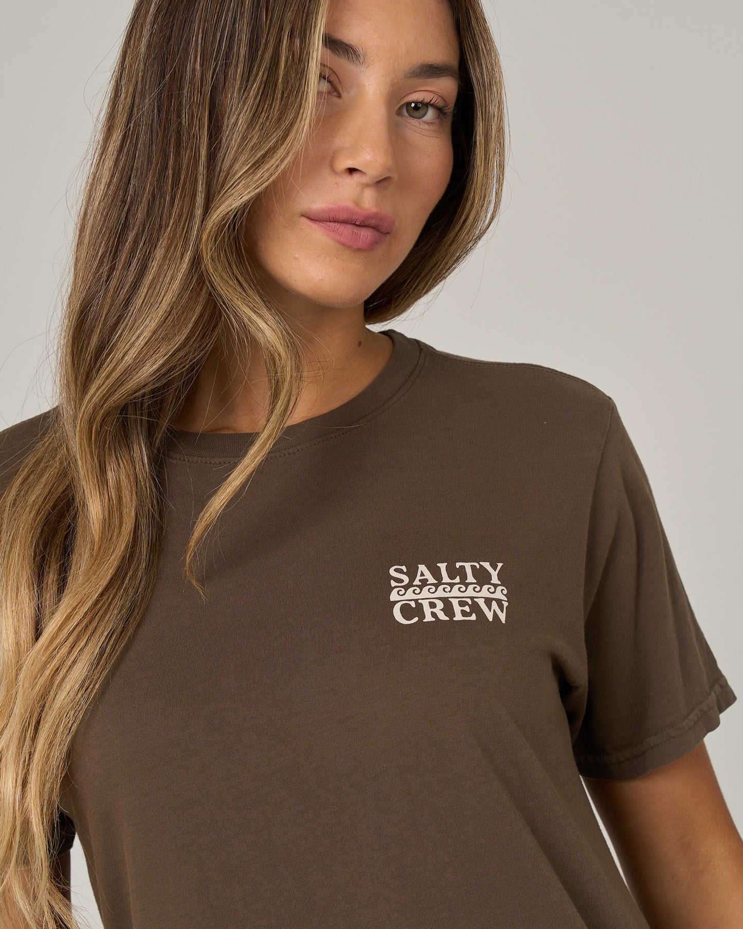 A woman with long, wavy blonde hair is wearing the Salty Crew Lobster Roll Boyfriend Tee in washed brown—a short sleeve cotton jersey t-shirt featuring white “Salty Crew” text on the chest—while standing against a neutral gray background.