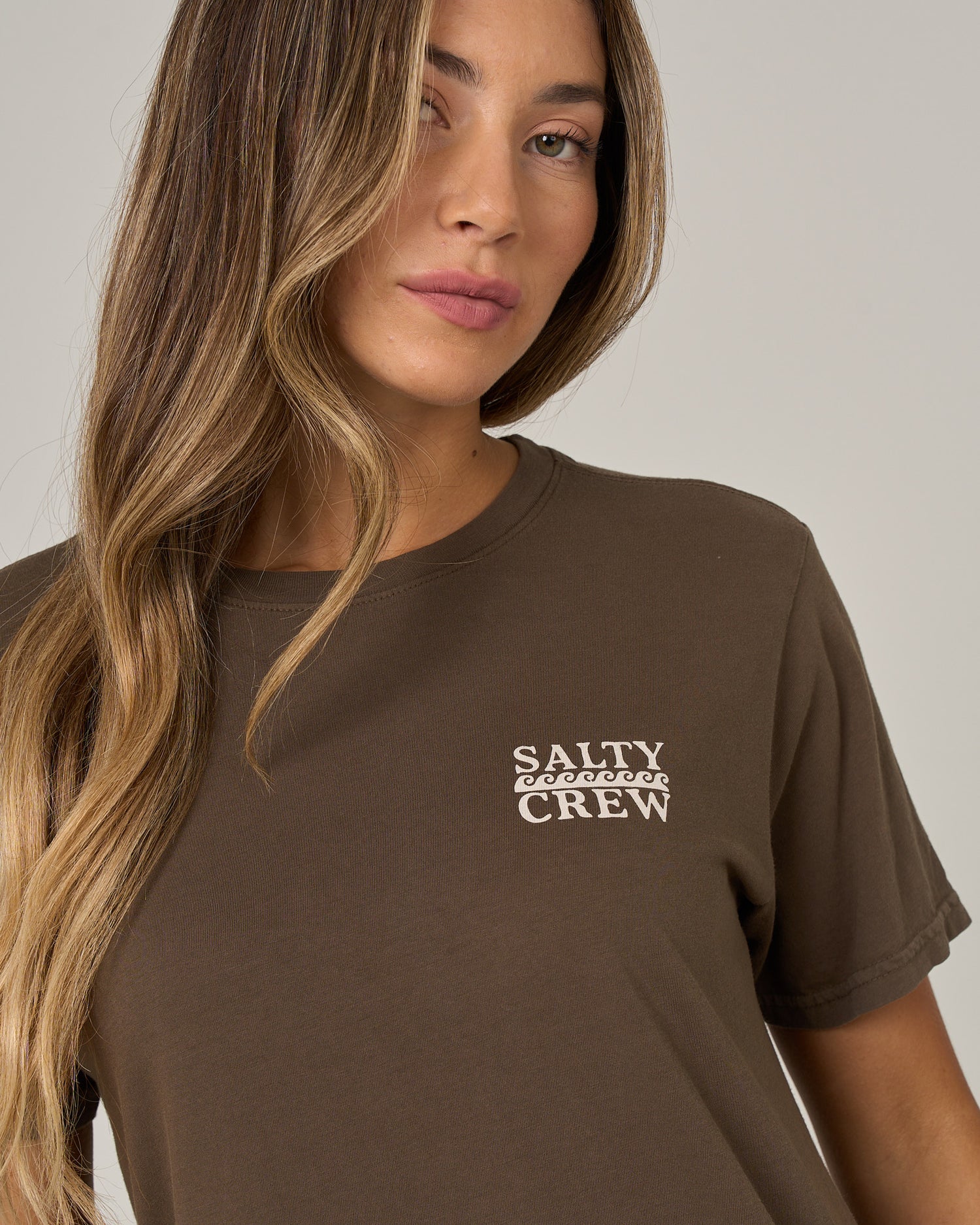 A woman with long, wavy blonde hair is wearing the Salty Crew Lobster Roll Boyfriend Tee in washed brown—a short sleeve cotton jersey t-shirt featuring white “Salty Crew” text on the chest—while standing against a neutral gray background.