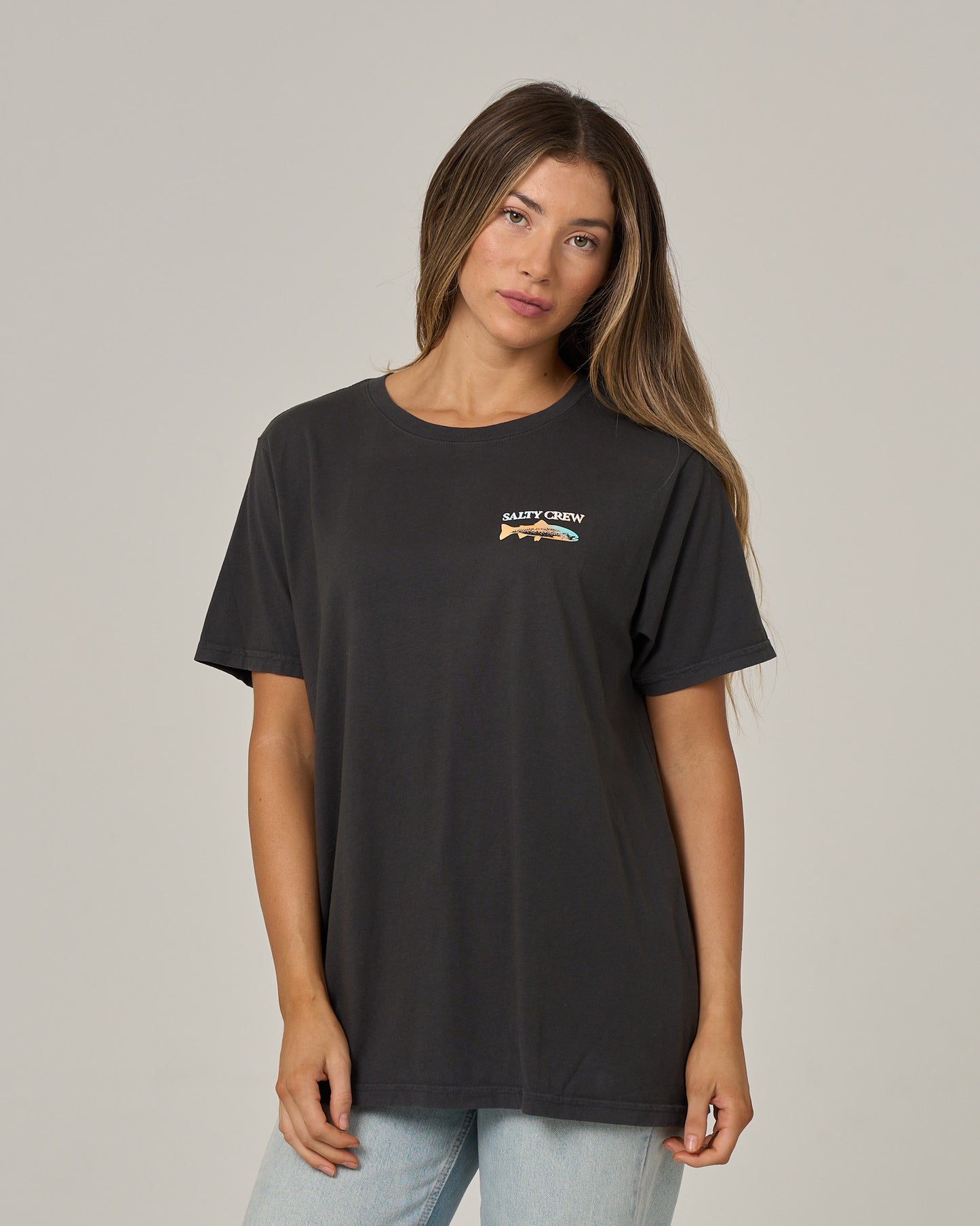 A woman with long brown hair is wearing the Salty Crew Daily Catch Boyfriend Tee in charcoal, a relaxed fit soft cotton jersey shirt with a small chest graphic, paired with light blue jeans and standing against a plain light gray background.