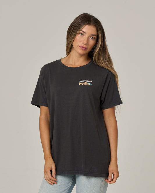 A woman with long brown hair is wearing the Salty Crew Daily Catch Boyfriend Tee in charcoal, a relaxed fit soft cotton jersey shirt with a small chest graphic, paired with light blue jeans and standing against a plain light gray background.