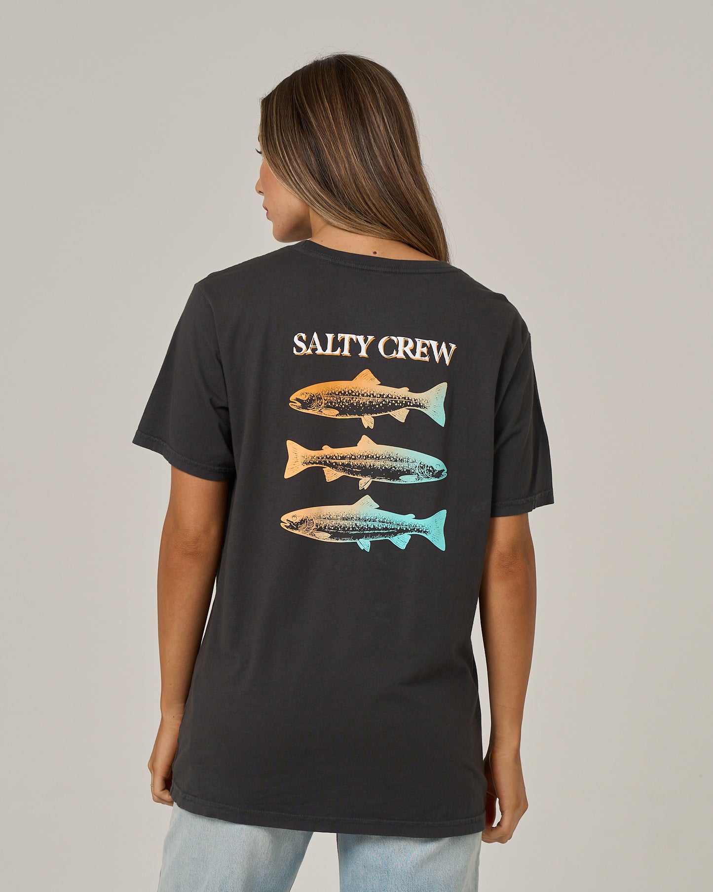 A woman is seen from behind wearing the Salty Crew Daily Catch Boyfriend Tee in charcoal, a relaxed fit cotton jersey t-shirt with short sleeves. The shirt features “SALTY CREW” above a vertical graphic of three trout, each illustrated in unique orange and blue gradient hues.
