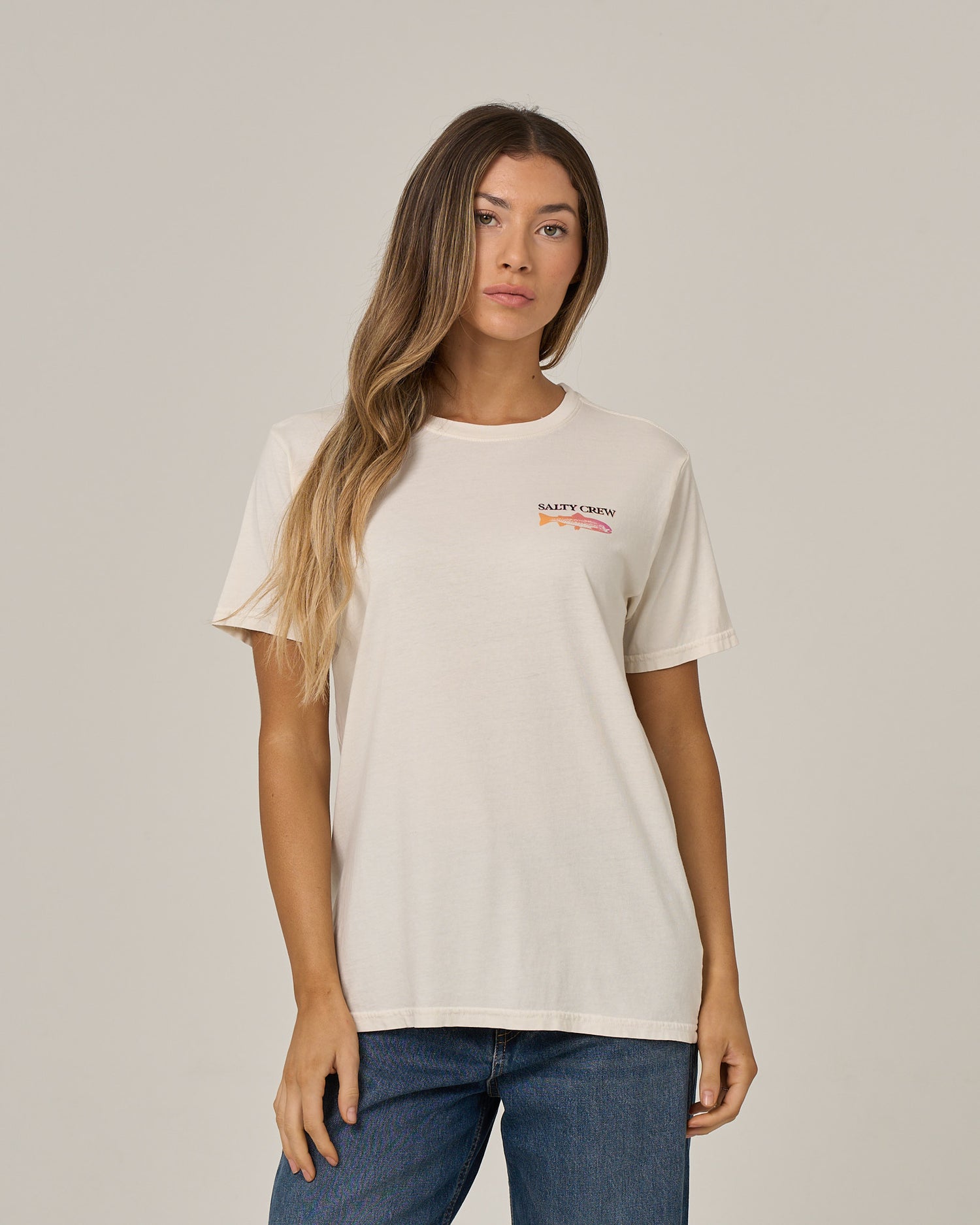 A woman with long, wavy hair wears the Salty Crew Daily Catch Boyfriend Tee in off white—a relaxed fit cotton jersey t-shirt with short sleeves and small text on the upper left side—paired with blue jeans, standing against a plain light background.