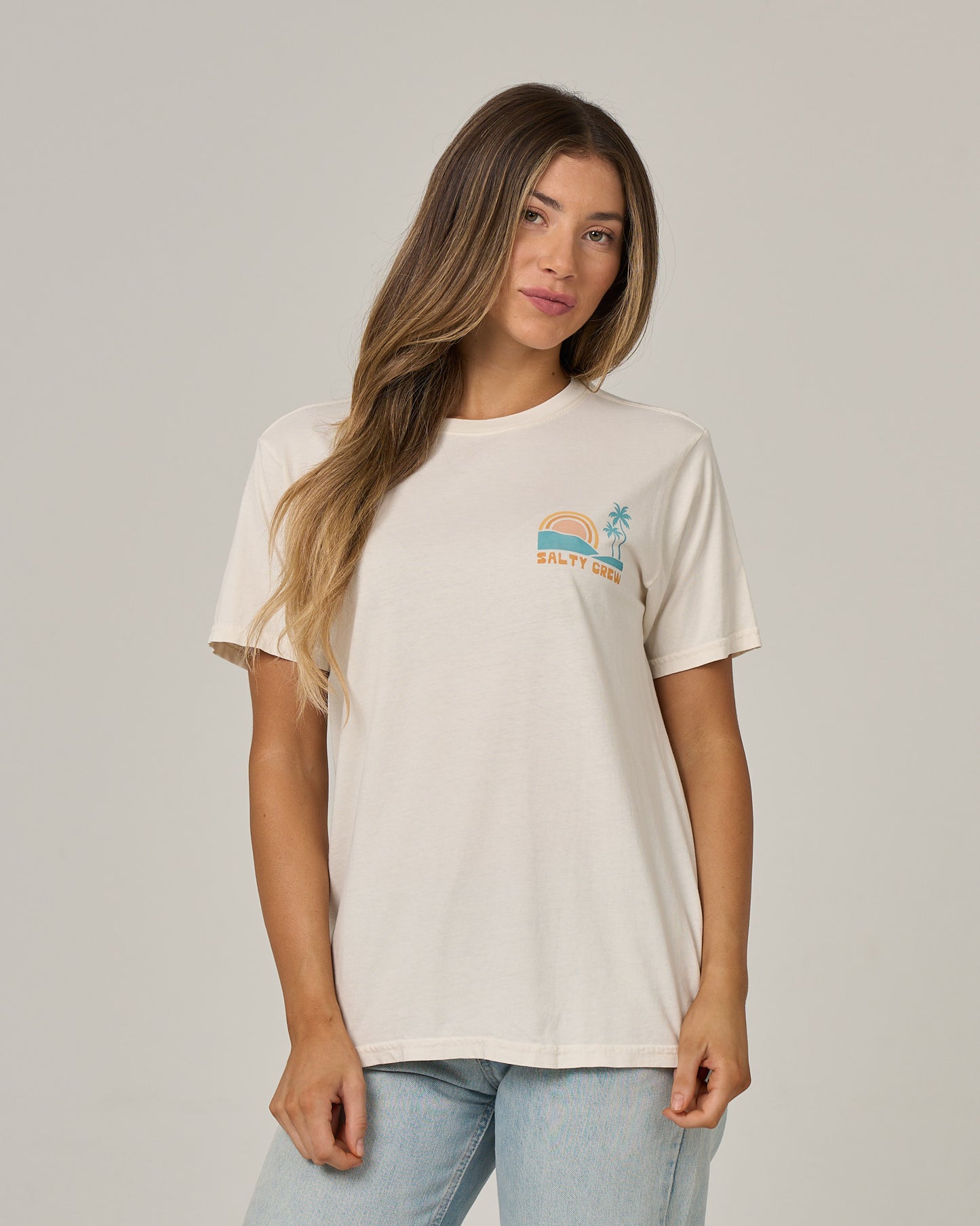 A woman wearing the Salty Crew Sushi Roll Boyfriend Tee in Off White—featuring a relaxed fit, short sleeves, and a small graphic with a rainbow and palm tree above the Salty Crew logo on the chest—stands against a plain background. This cotton jersey t-shirt offers effortless style and comfort for any casual look.