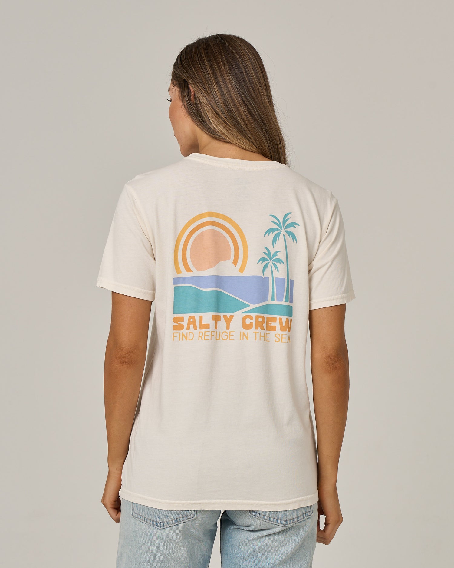 A woman with long hair is seen from behind wearing the Salty Crew Sushi Roll Boyfriend Tee in off white, a relaxed fit cotton jersey shirt. The back features a graphic of a sun, ocean waves, and palm trees, along with the words “SALTY CREW FIND REFUGE IN THE SEA.”.