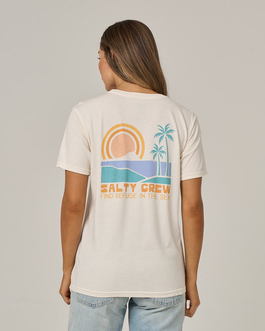 A woman with long hair is seen from behind wearing the Salty Crew Sushi Roll Boyfriend Tee in off white, a relaxed fit cotton jersey shirt. The back features a graphic of a sun, ocean waves, and palm trees, along with the words “SALTY CREW FIND REFUGE IN THE SEA.”.
