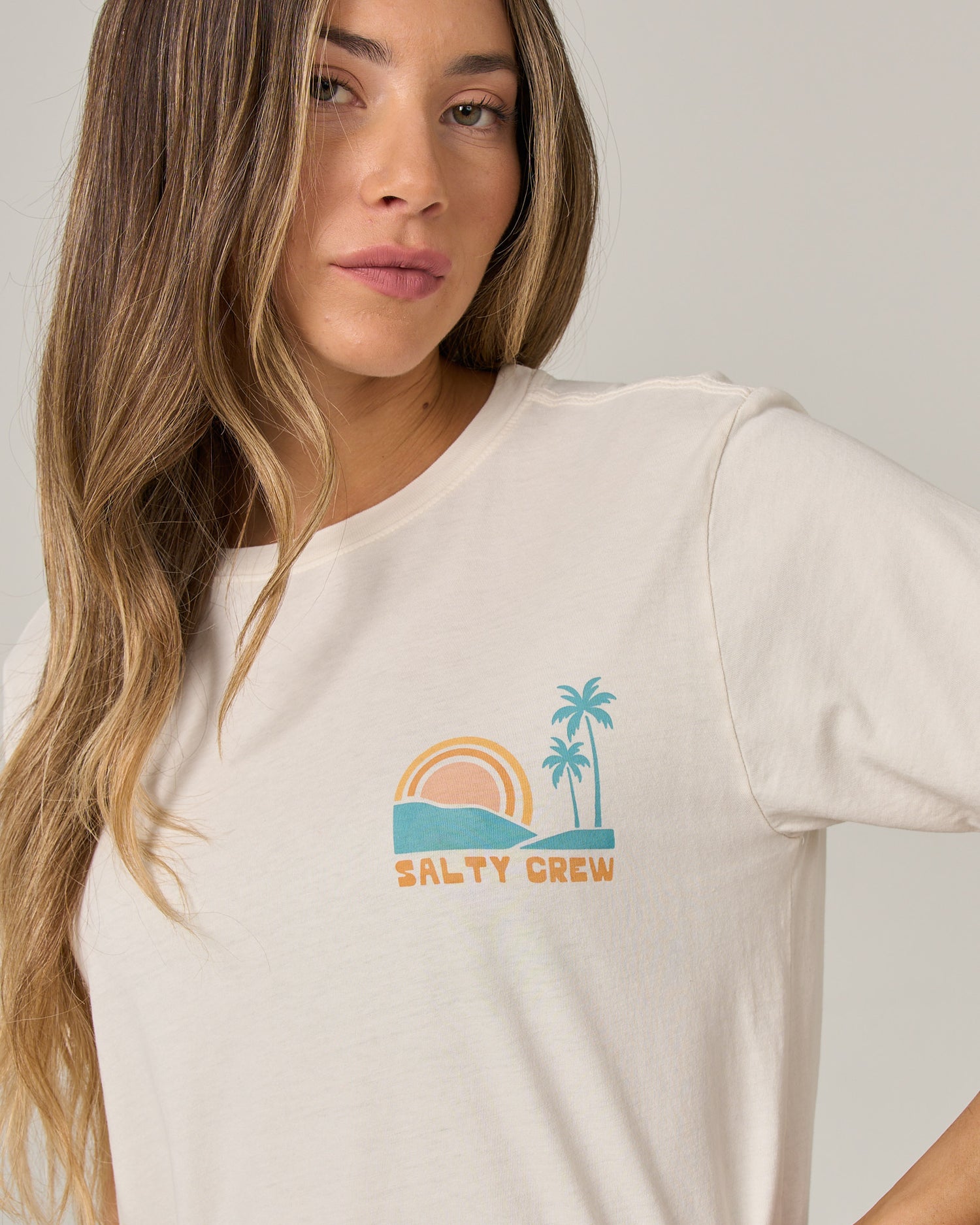 A woman with long, wavy hair wears the Salty Crew Sushi Roll Boyfriend Tee in off white—a relaxed fit, soft cotton jersey T-shirt featuring a small graphic of a sunset, ocean, and palm trees with the words “Salty Crew” on the left chest.