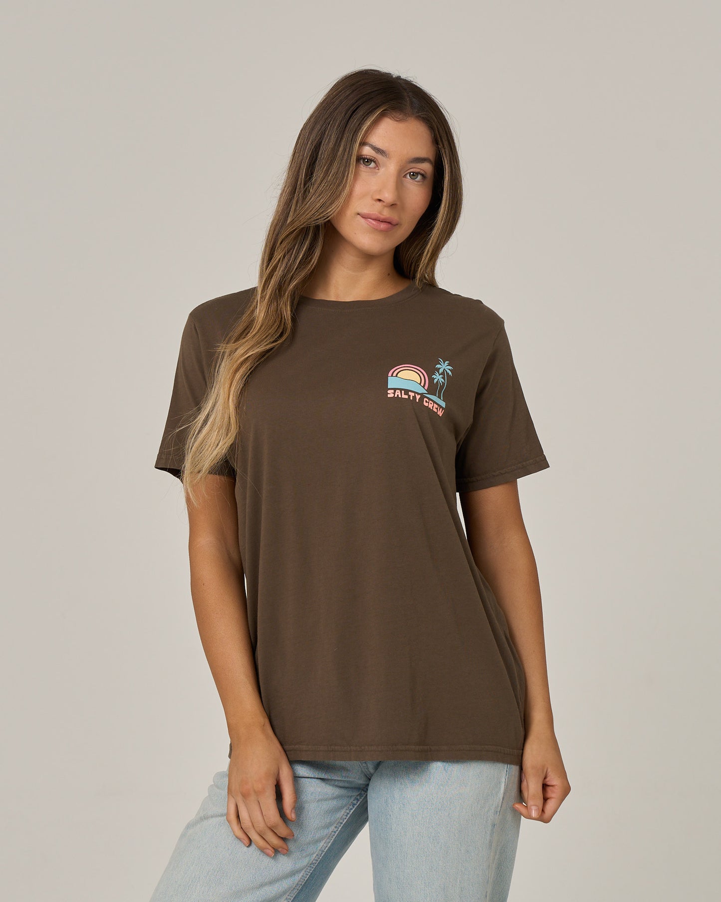 A woman with long hair wears the Salty Crew Sushi Roll Boyfriend Tee in washed brown, a short-sleeve cotton jersey featuring a small rainbow, palm tree, and Salty Crew logo on the front. She pairs it with light blue jeans and stands against a plain, light background.