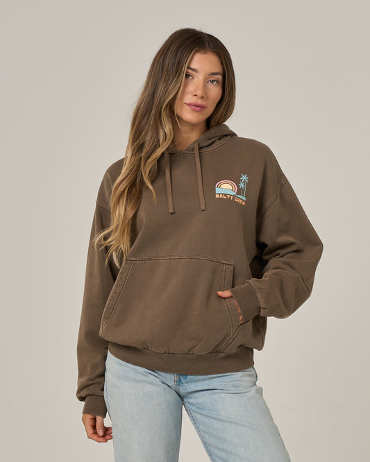 A woman with long wavy hair wears the Salty Crew Sushi Roll Hoodie in washed brown, featuring a rainbow and palm tree design on the chest, a cozy kangaroo pocket, and soft cotton fleece construction. She pairs it with light blue jeans while standing against a plain background.