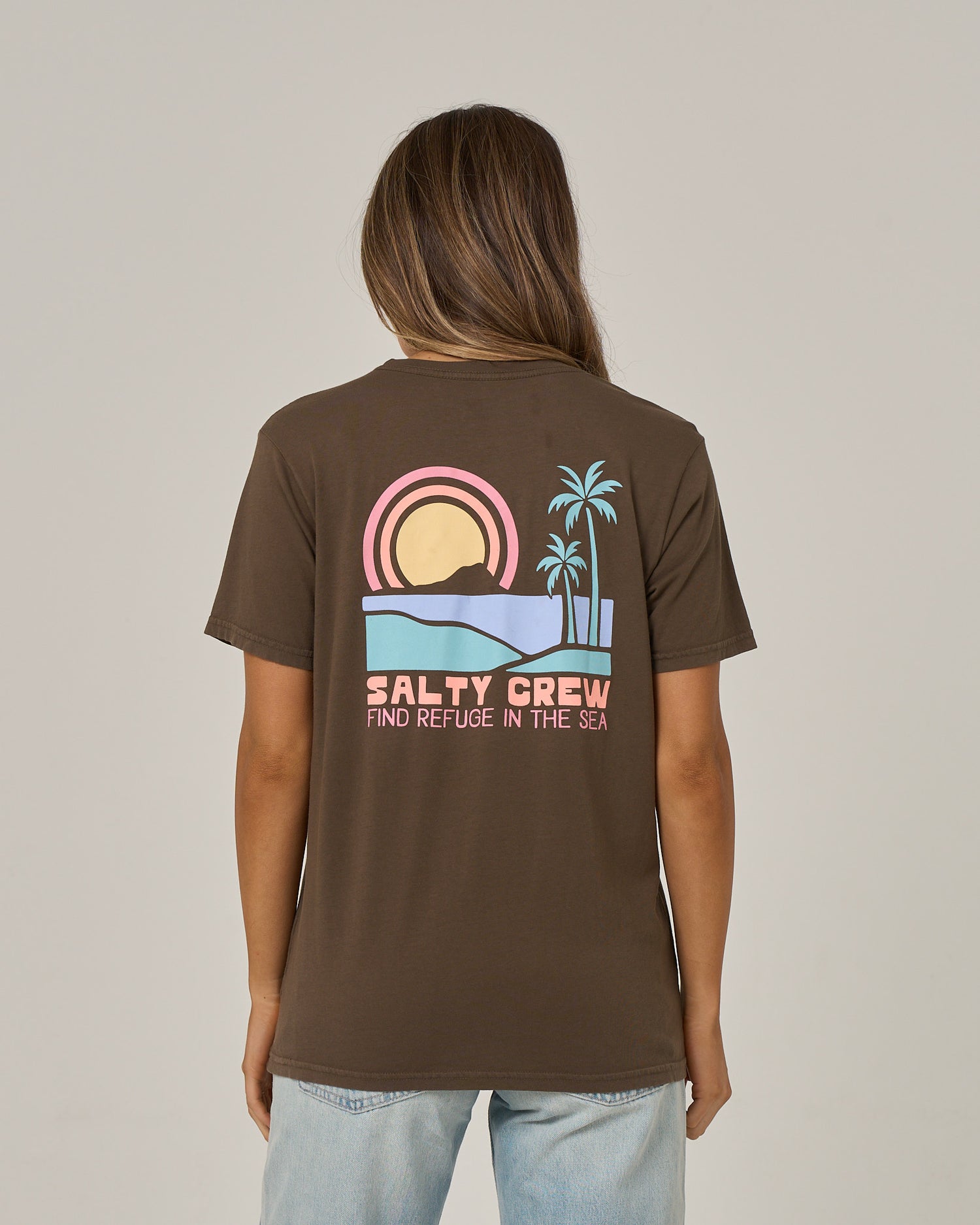 A person is seen from behind wearing the Salty Crew Sushi Roll Boyfriend Tee in washed brown, a relaxed fit short sleeve crafted from soft cotton jersey. The tee features a graphic of a sunset, waves, and palm trees along with the text “SALTY CREW FIND REFUGE IN THE SEA.”.