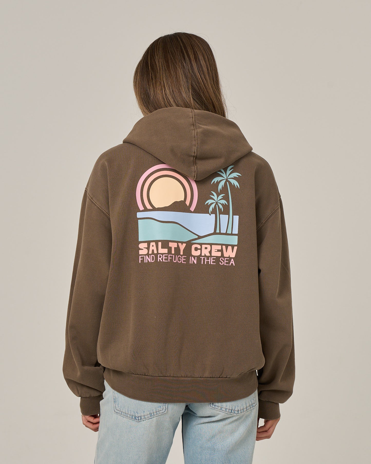 A person is seen from behind wearing the Salty Crew Sushi Roll Hoodie in washed brown, featuring a bold back graphic of a sunset, ocean, palm trees, and the phrase “Find Refuge in the Sea.” This oversized hoodie is crafted from soft cotton fleece and comes equipped with a classic kangaroo pocket.