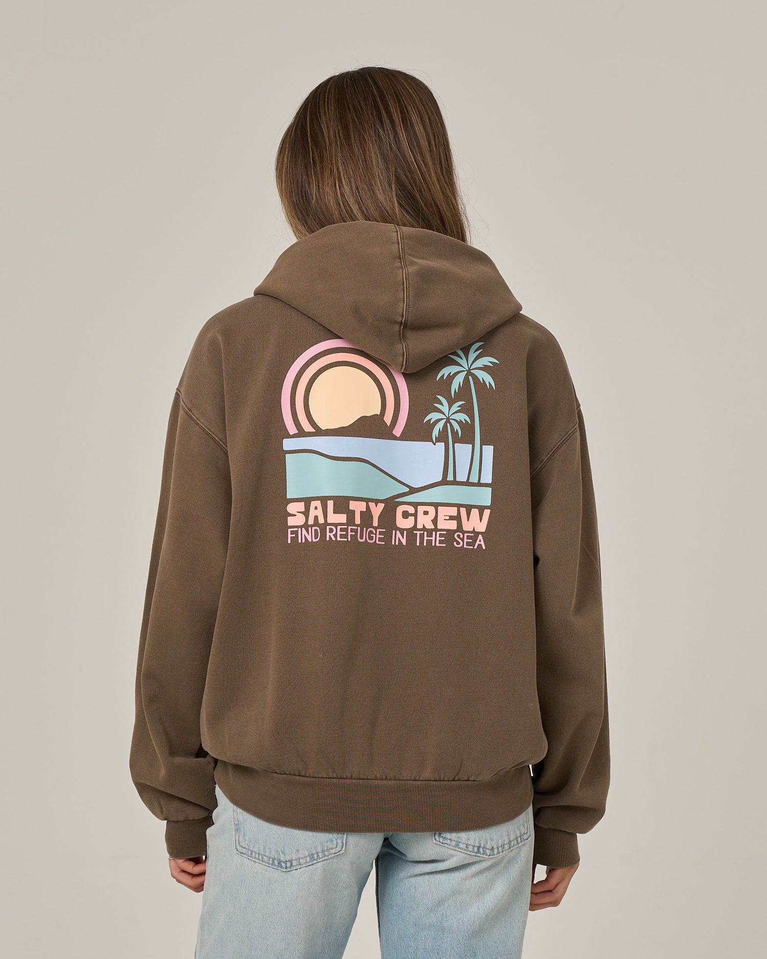 A person is seen from behind wearing the Salty Crew Sushi Roll Hoodie in washed brown, featuring a bold back graphic of a sunset, ocean, palm trees, and the phrase “Find Refuge in the Sea.” This oversized hoodie is crafted from soft cotton fleece and comes equipped with a classic kangaroo pocket.