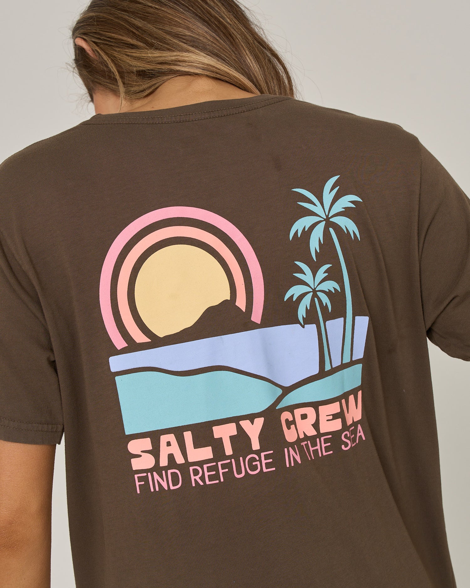 Wearing the Salty Crew Sushi Roll Boyfriend Tee in washed brown—a relaxed fit, short-sleeve top made from soft cotton jersey—this piece showcases a graphic of a sunset, palm trees, mountains, and pastel “SALTY CREW FIND REFUGE IN THE SEA” text on the back.