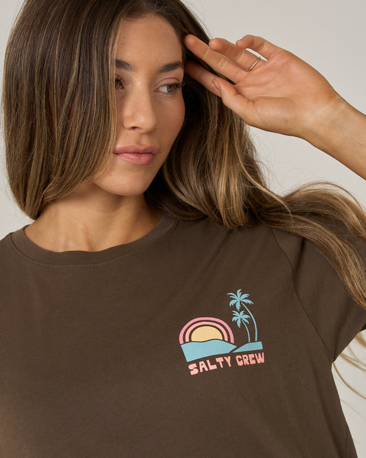 A woman with long hair wears the Salty Crew Sushi Roll Boyfriend Tee in washed brown, which features a pastel graphic of a sun, ocean waves, two palm trees, and the words SALTY CREW. She looks to the side with her hand raised near her head.