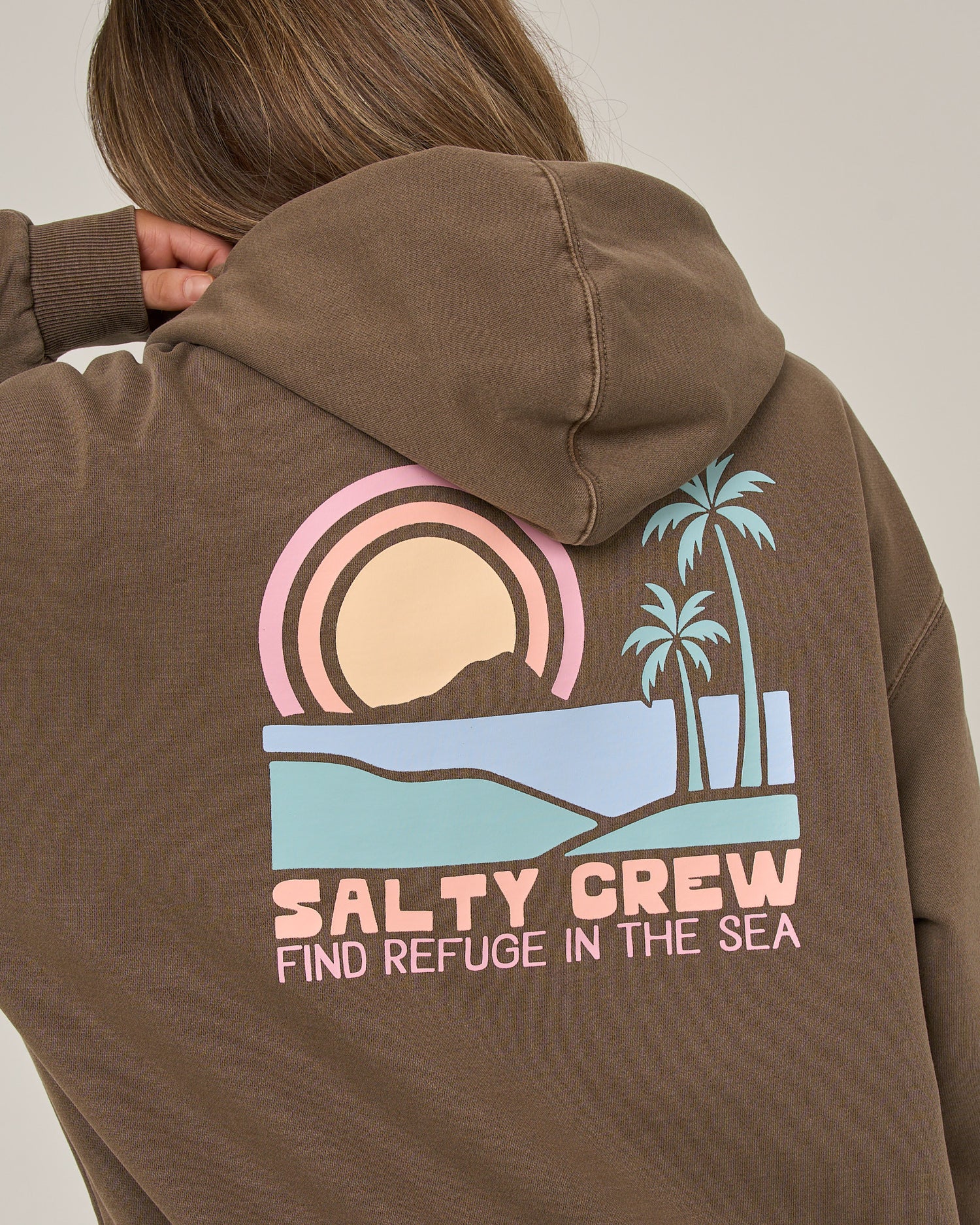 A person wears the Salty Crew Sushi Roll Hoodie in washed brown, crafted from soft cotton fleece. The back showcases a pastel-colored graphic featuring a sunset, palm trees, and the phrase “Salty Crew Find Refuge in the Sea.” A classic kangaroo pocket adds to this laidback style.