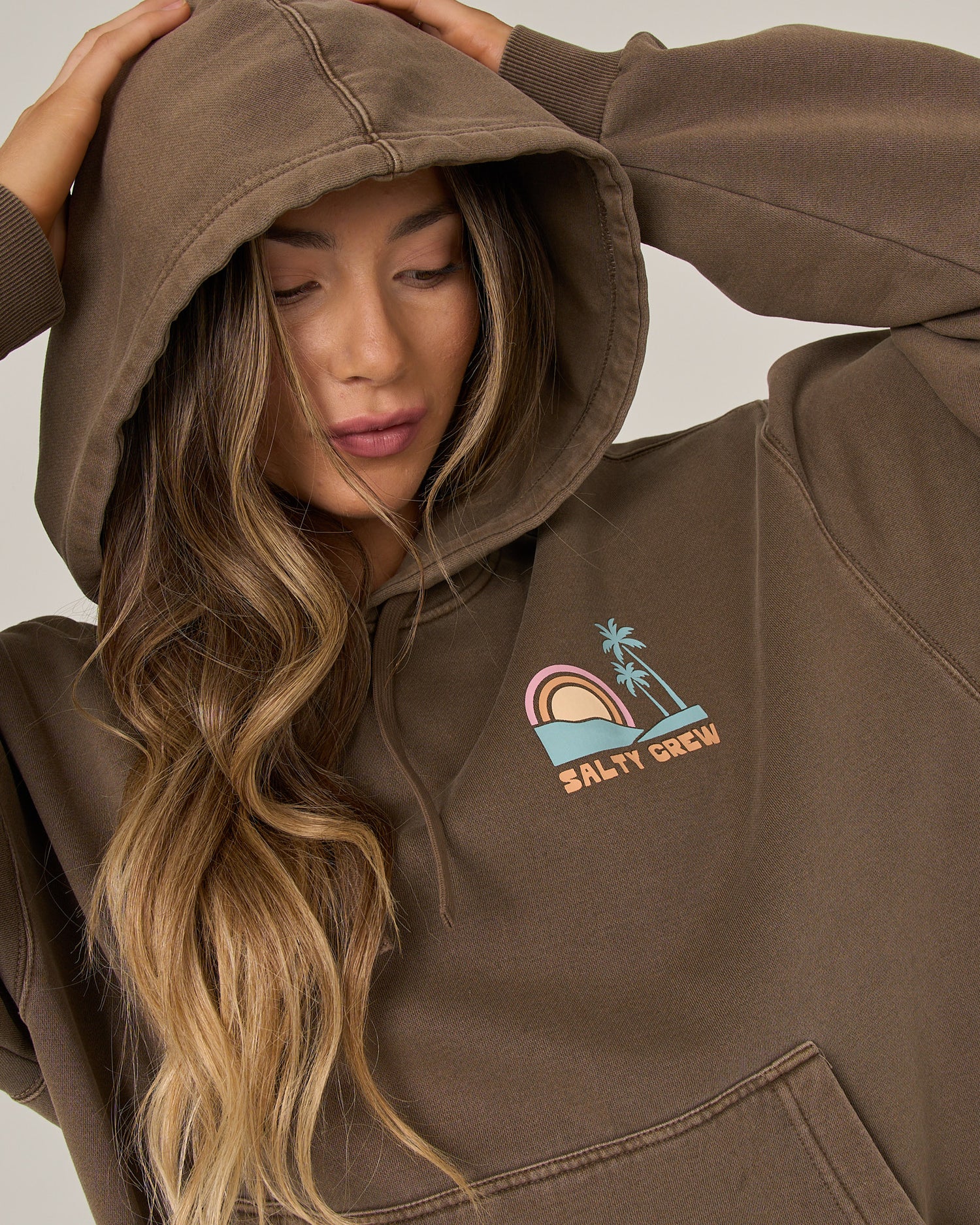 A person with long wavy hair, hood up and looking down, wears the Salty Crew Sushi Roll Hoodie in washed brown—an oversized cotton fleece hoodie featuring a rainbow and palm tree design along with the Salty Crew logo, plus a roomy kangaroo pocket for extra comfort.