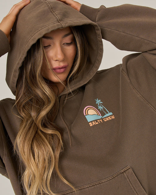 A person with long wavy hair, hood up and looking down, wears the Salty Crew Sushi Roll Hoodie in washed brown—an oversized cotton fleece hoodie featuring a rainbow and palm tree design along with the Salty Crew logo, plus a roomy kangaroo pocket for extra comfort.
