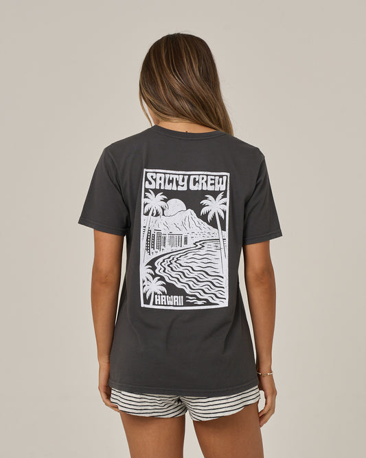 A person with long hair faces away, wearing the Salty Crew Aloha Breeze Boyfriend Tee in charcoal—a relaxed fit shirt crafted from soft cotton jersey and adorned on the back with a white tropical graphic featuring palm trees, mountains, and waves. Designed with short sleeves, this tee pairs effortlessly with striped shorts against a plain, light background.