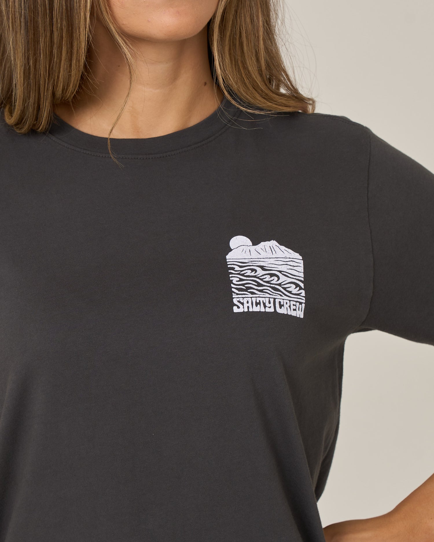 Wearing the Salty Crew Aloha Breeze Boyfriend Tee in charcoal, this relaxed fit cotton jersey shirt features a white graphic of waves, a sun, mountains, and “SALTY CREW” text on the upper chest.