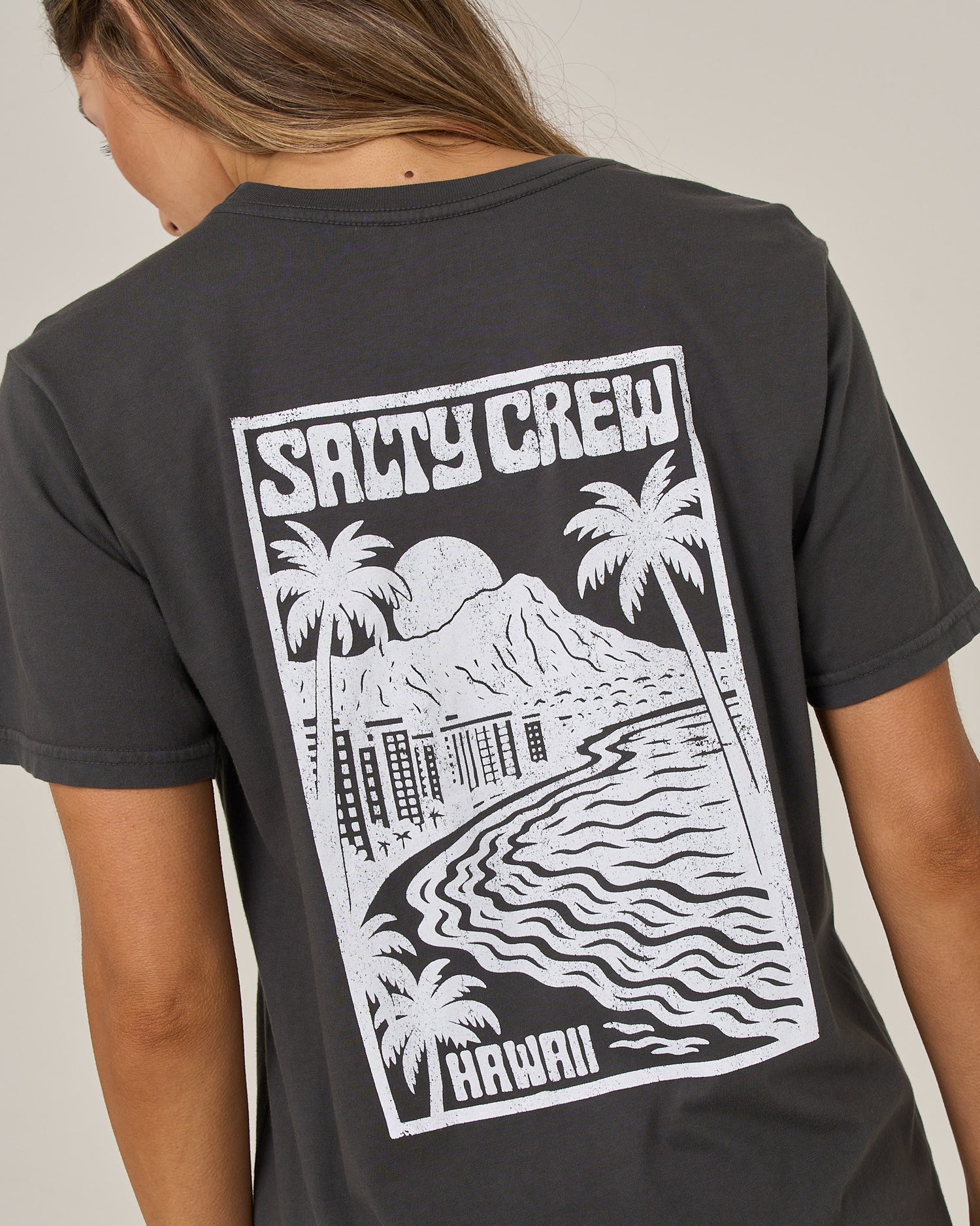 A woman with long hair wears the Salty Crew Aloha Breeze Boyfriend Tee in charcoal, a relaxed fit short sleeve style crafted from soft cotton jersey. The back features a white graphic of palm trees, mountains, water, and buildings along with the words “SALTY CREW” and “HAWAII.”.